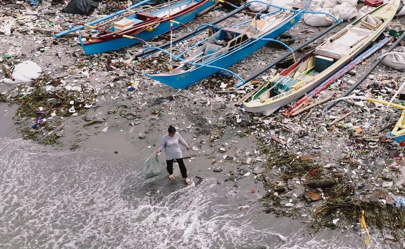 A collector gathering plastic from a shoreline beside fishing boats in the Philippines